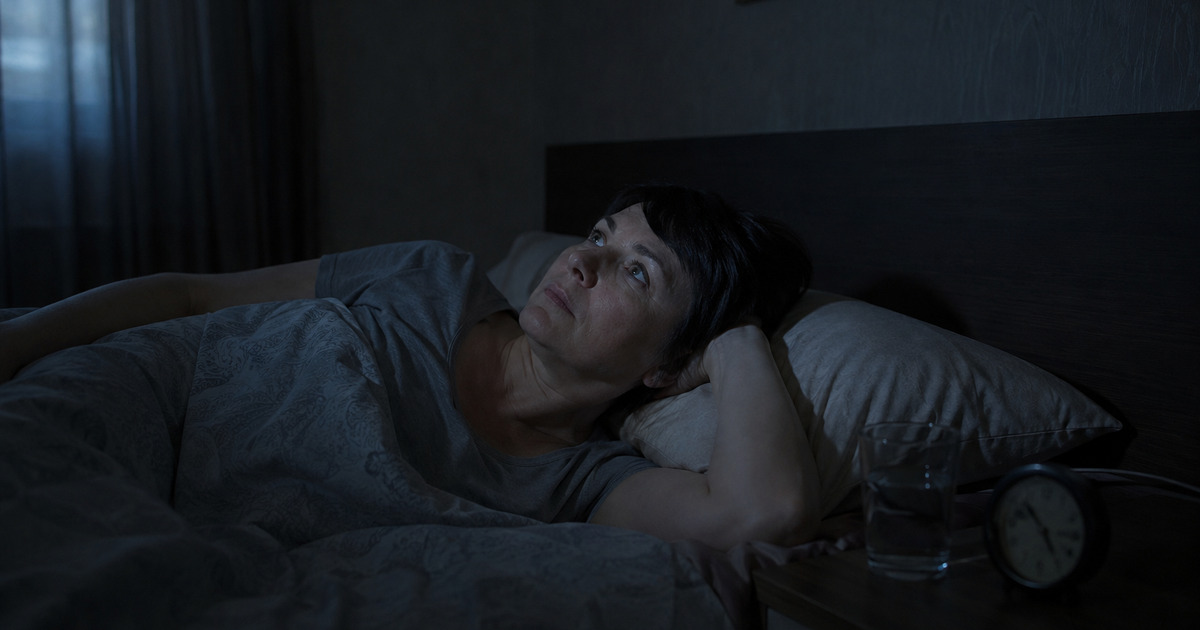 женщина 45+ лежит ночью в постели при выключенном свете, глаза открыты, за окном тёмное небо